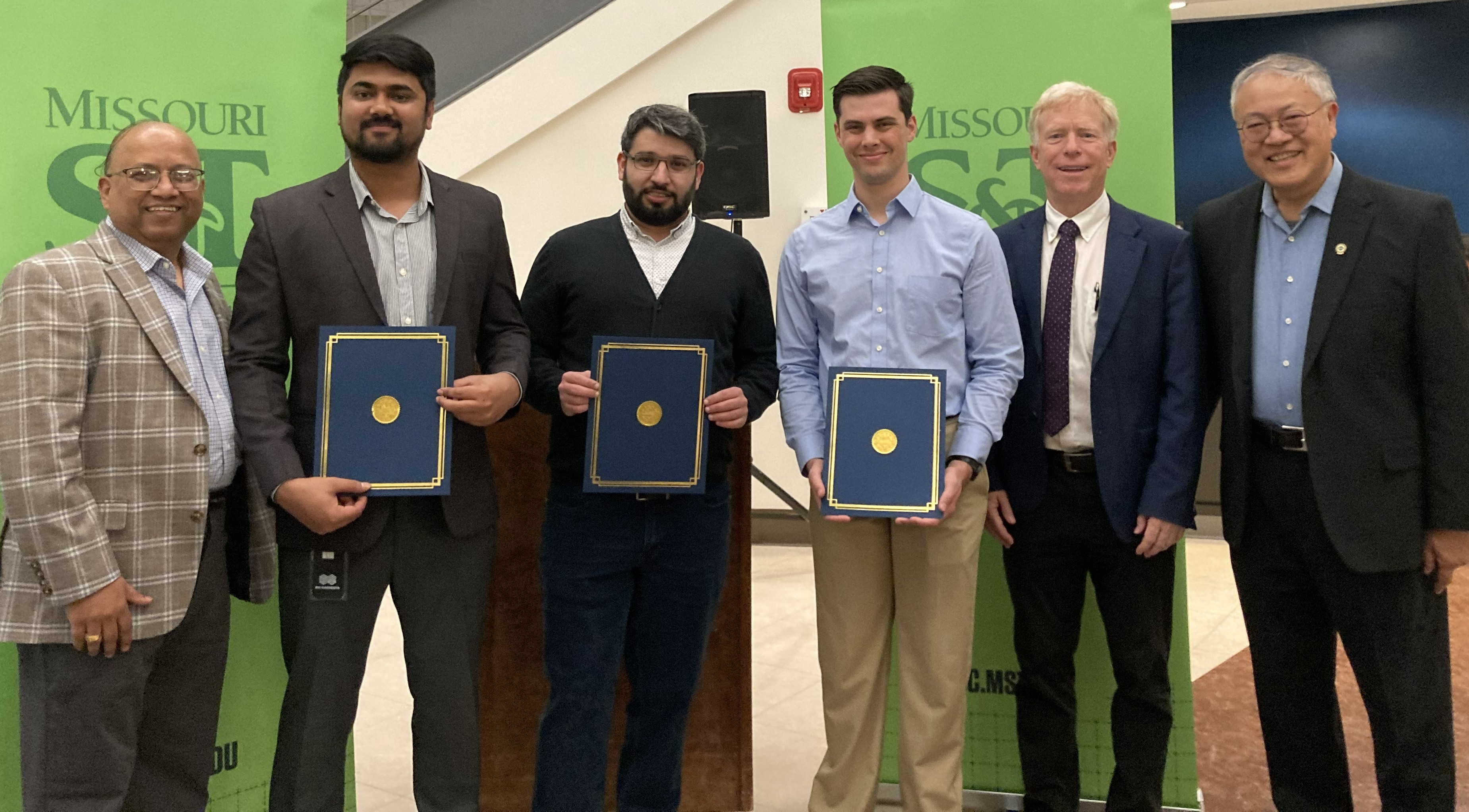 A group of six men stand indoors, smiling. Three hold certificates. Behind them are green Missouri S&T banners. The mood is celebratory and formal.